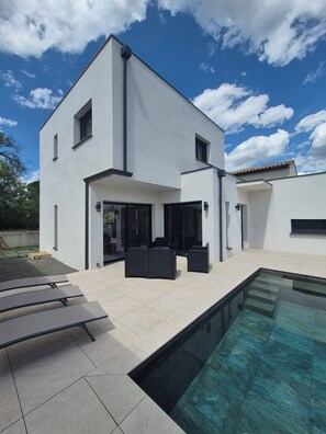 Ein modernes, zweistöckiges Haus mit Swimmingpool, Sitzgelegenheiten im Freien und blauem Himmel.