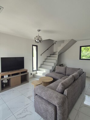 Ein modernes Wohnzimmer mit einem großen Flachbildfernseher, einem grauen Ecksofa, einer Holz-Unterhaltungseinrichtung, einer Treppe und einem runden Couchtisch.