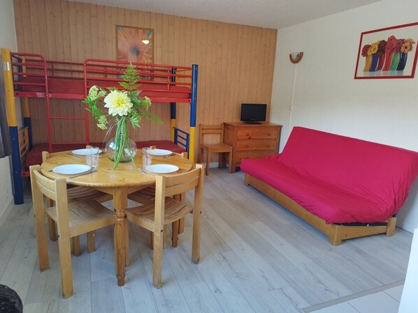 Ein gemütlicher Raum mit Esstisch für zwei Personen, einem roten Sofa, einem Fernseher und einem Etagenbett.