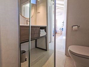 Un baño con inodoro, lavamanos con espejo, toallero y cubo de basura.
