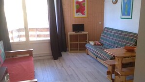 Ett litet, ombonat rum med trägolv, en röd soffa, ett träbord, en tv på ett stativ och ett fönster med gardiner.