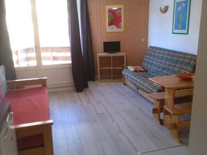 Ein kleiner, gemütlicher Raum mit Holzboden, einem roten Sofa, einem Holztisch, einem Fernseher auf einem Schrank und einem Fenster mit Vorhängen.