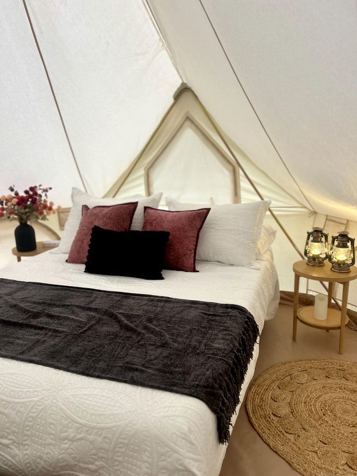 A tent-style room with a bed, pillows, a blanket, a small table with lanterns, and a vase with flowers.