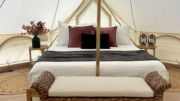 A bed in a tent with a canopy, a wicker chair, a small table with a vase of flowers, and a lantern on a bedside table.