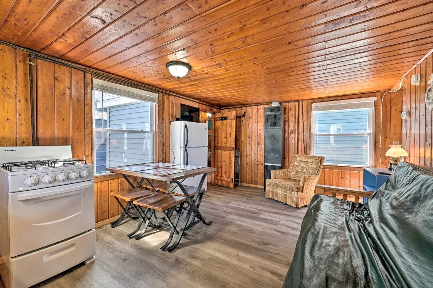 A cabin interior with a wooden ceiling, a Danby stove, a refrigerator, a dining table with benches, a chair, and a sofa.