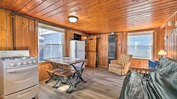 A cabin interior with a wooden ceiling, a Danby stove, a refrigerator, a dining table with benches, a chair, and a sofa.