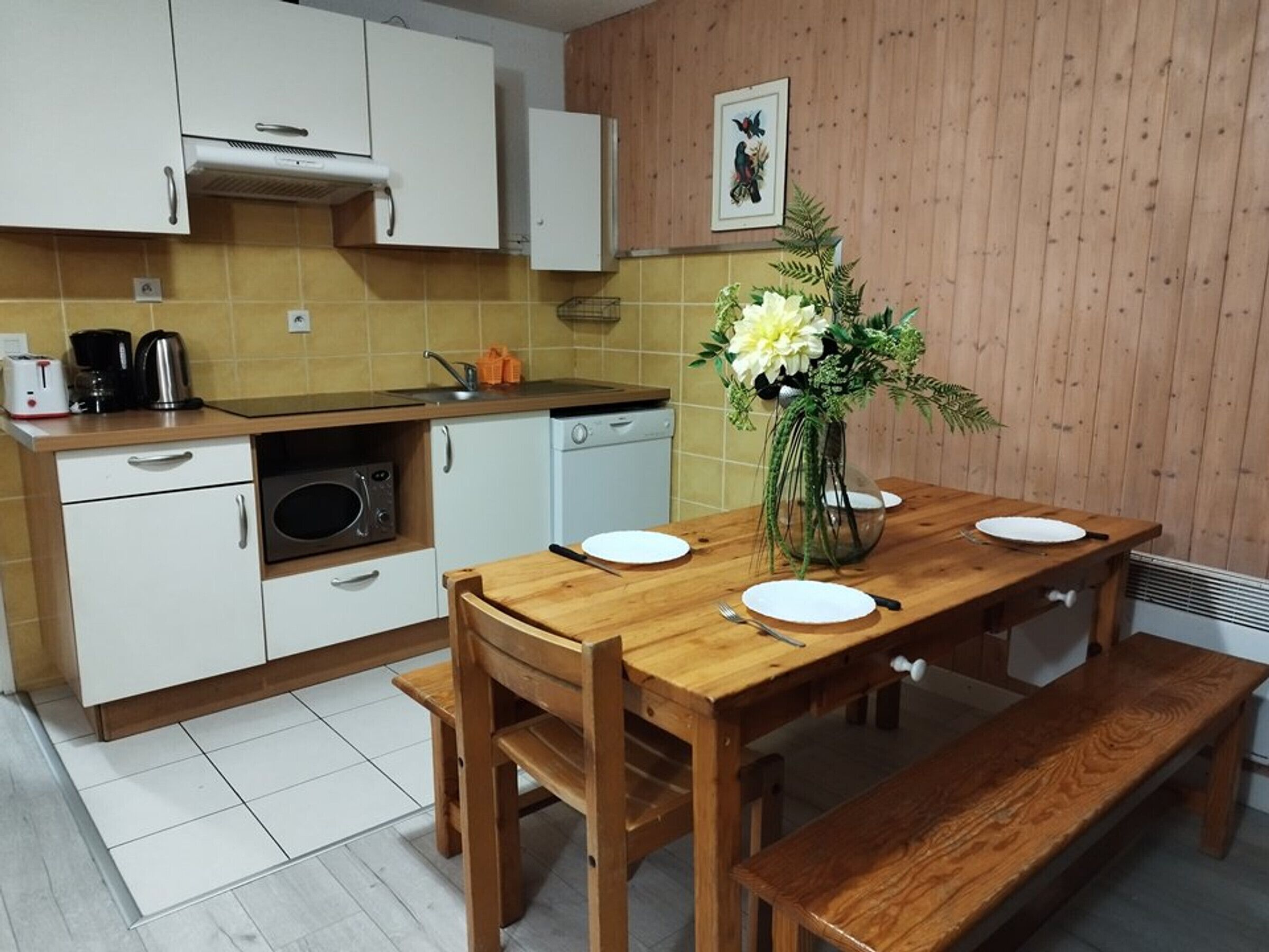 Une cuisine compacte avec des meubles blancs, un four à micro-ondes et un évier. On y trouve une table à manger en bois, avec de l’assiette blanche et un vase de fleurs.