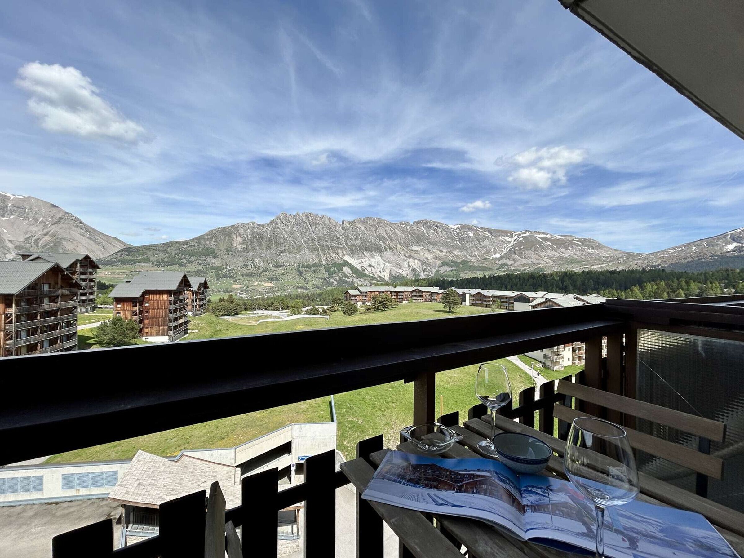 Un balcón con vistas a montañas, una mesa de madera con una copa de vino y un folleto.