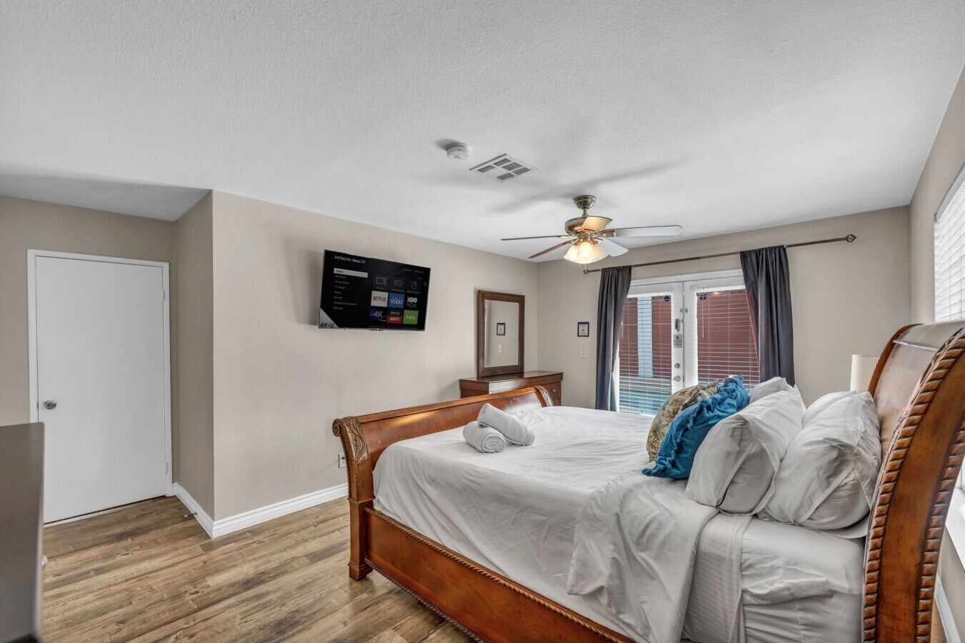 A bedroom with a wooden bed, a ceiling fan, a TV mounted on the wall, and a window with curtains.