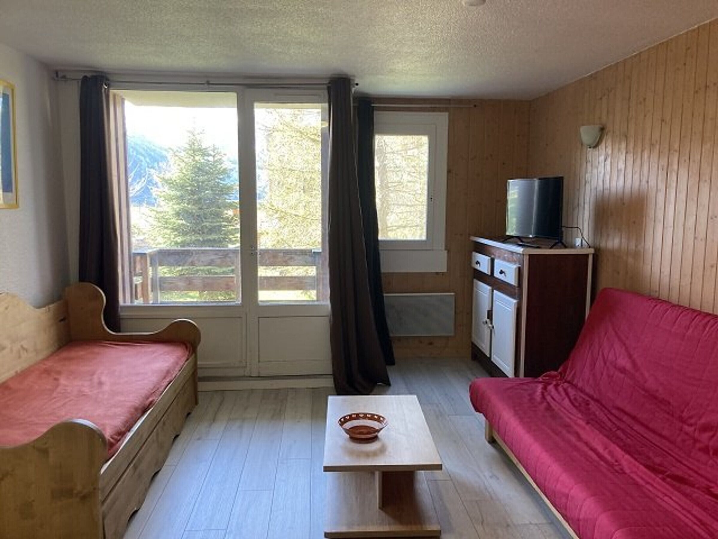 A room with a wooden floor, a red sofa, a wooden bench, a small table, a television on a cabinet, and a sliding glass door with curtains.