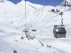 Ein Sesselliftstuhl, der an Seilen hängt, mit verschneiter Berglandschaft im Hintergrund.
