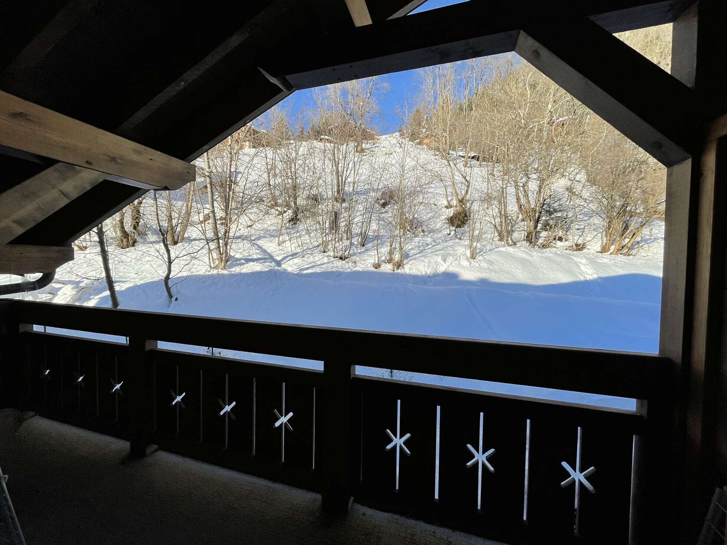 A snow-covered landscape with bare trees, viewed through a wooden structure with a black railing and star-shaped cutouts.