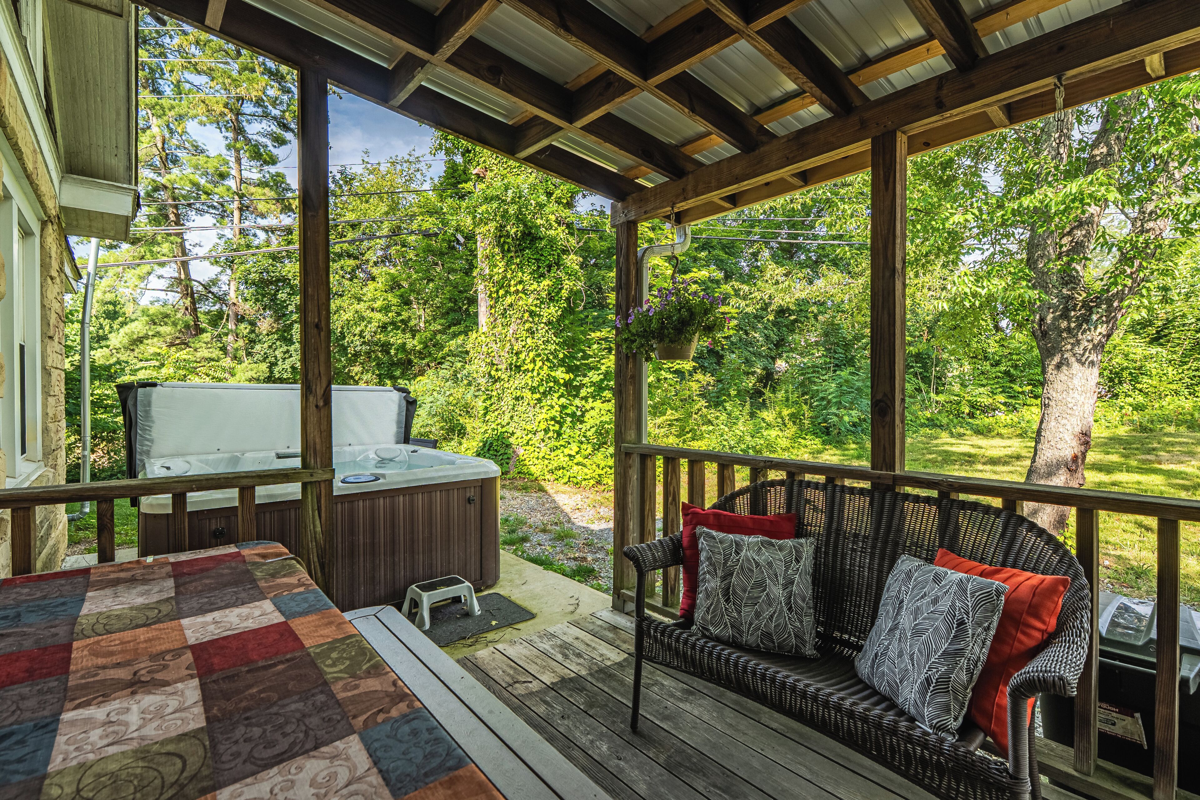 A covered outdoor deck with a sofa, a hot tub, and a view of a lush garden.