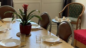 Ein Esszimmertisch ist gedeckt mit Tellern, Gläsern und Besteck, daneben ein Blumentopf und ein Stuhl mit einem floralen Kissen.