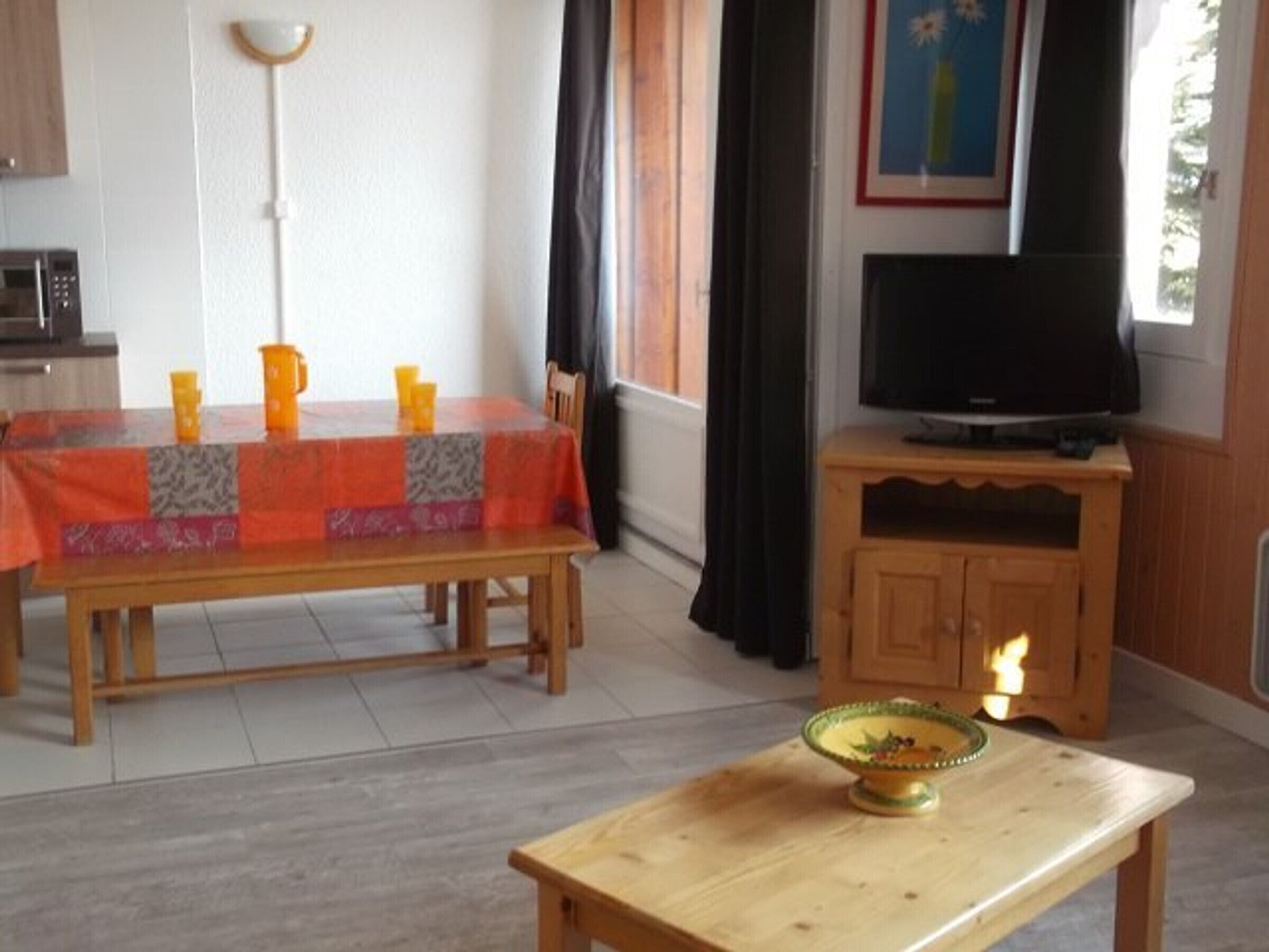 Un coin repas avec une table recouverte d’une nappe à motifs, un téléviseur posé sur un meuble en bois et une petite table sur laquelle se trouve un bol.