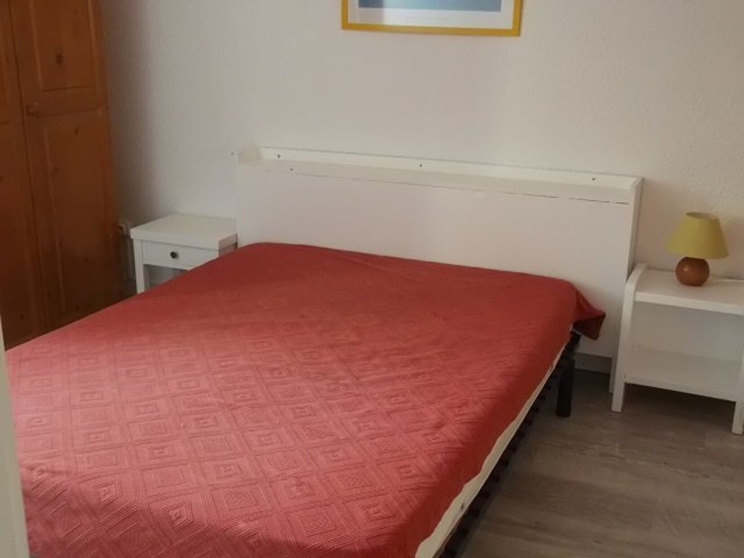 A bedroom with a red patterned bedspread, a white headboard, a bedside table with a lamp, and a wooden wardrobe.