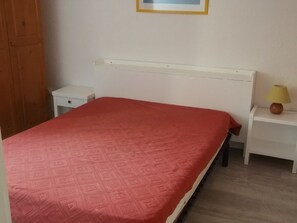 A bedroom with a red patterned bedspread, a white headboard, a bedside table with a lamp, and a wooden wardrobe. - Bright apartment near slopes - 8 guests (Orcières Merlette)