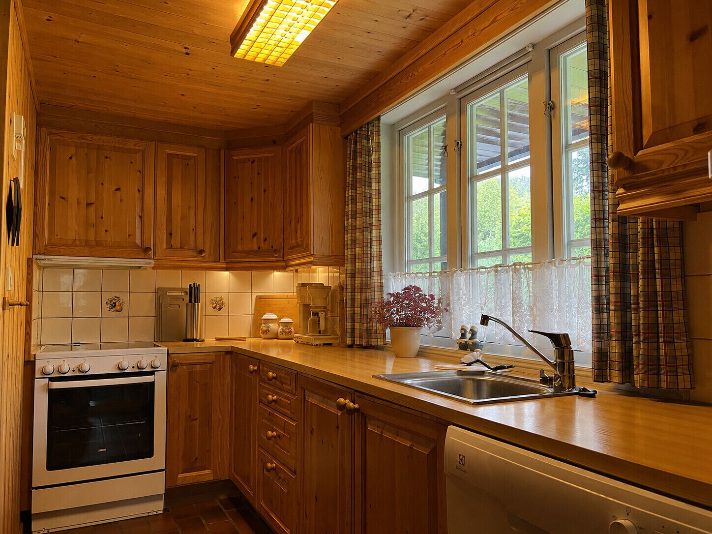Una cocina de madera con horno empotrado, pileta de acero inoxidable y ventana con cortinas.