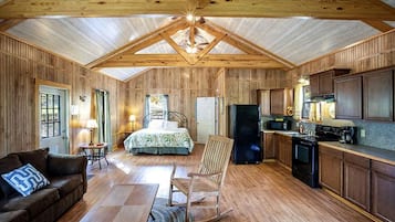 Interior de una cabaña con piso de madera, una cama, una mecedora, una zona de cocina y una mesa de comedor.