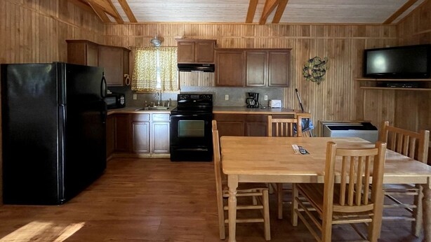 A compact kitchen with wooden cabinets, a black refrigerator, a stove, and a dining area with wooden chairs and a table. - Charming Pondview Cabin with Resort Amenities near Zwolle, Louisiana (Zwolle)