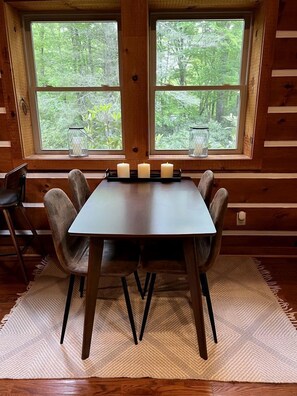A wooden dining table with four chairs, candles, and a view of a forest through a large window.