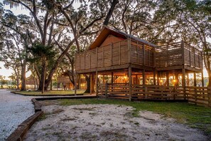 A wooden cabin with a covered porch, surrounded by trees and a gravel path.