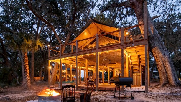 A wooden cabin with a covered deck, outdoor seating, and a fire pit. - Unique Marshfront Treehouse Stay with Heated Soaking Tub & Adventure-Inspired Escape in Townsend, Georgia (Townsend)