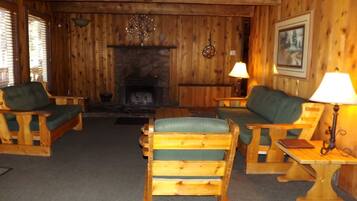 L’intérieur d’un chalet accueillant, avec des murs en bois, une cheminée, deux fauteuils en bois dotés de coussins verts et une petite table avec une lampe.