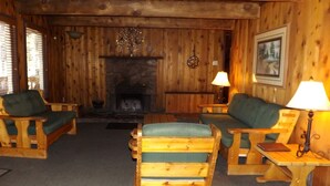 A cozy cabin interior with wooden walls, a fireplace, two wooden chairs with green cushions, and a small table with a lamp. - Spacious Cabin with Large Porch Ideal for Groups in Arizona (Flagstaff)