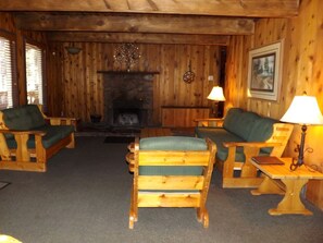 A cozy cabin interior with wooden walls, a fireplace, two wooden chairs with green cushions, and a small table with a lamp. - Spacious Cabin with Large Porch Ideal for Groups in Arizona (Flagstaff)