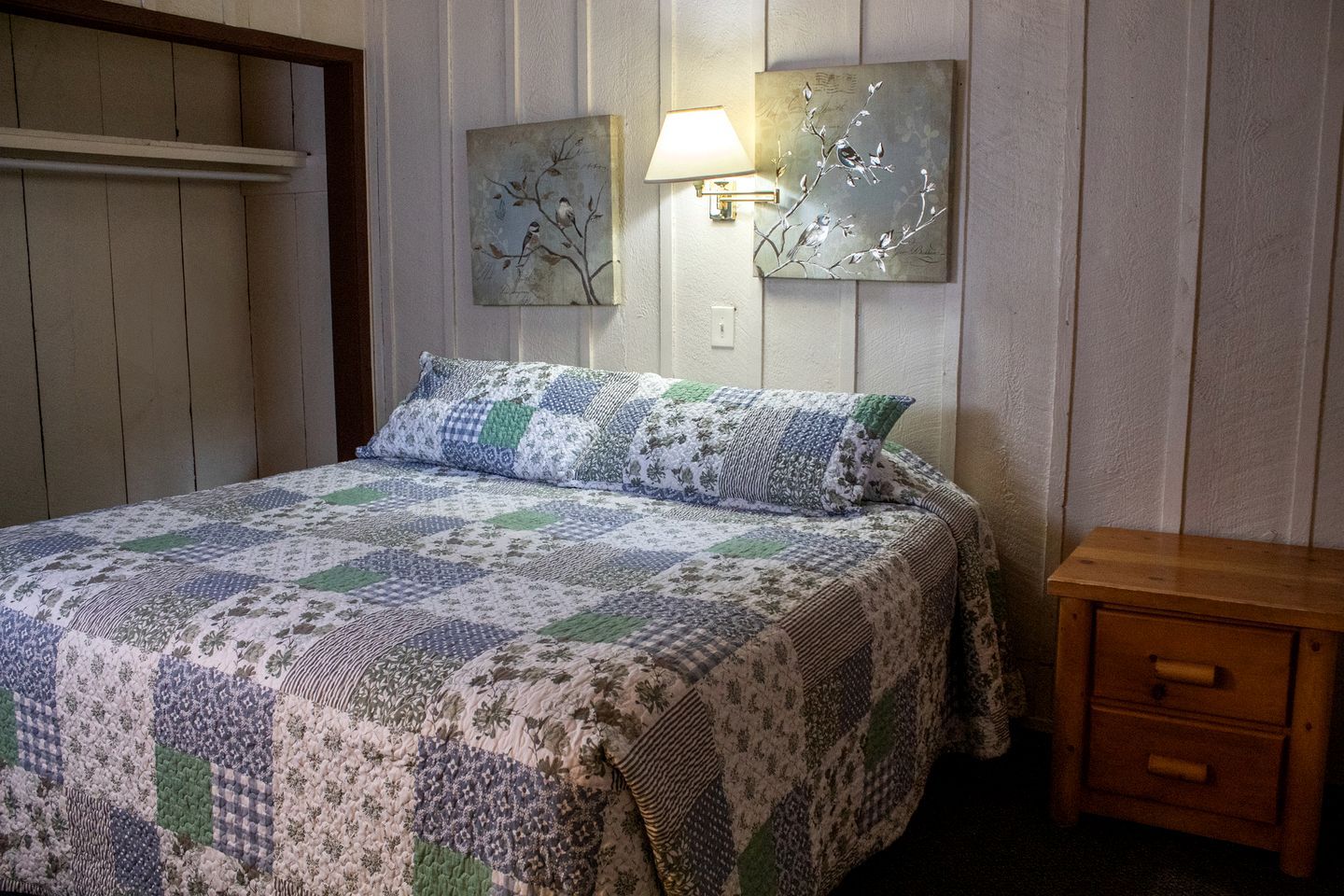 A bedroom with a bed, a nightstand, a wall-mounted lamp, and two wall art pieces.