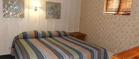 Ein Schlafzimmer mit gestreifter Bettdecke, zwei Wandlampen, einem Fenster mit Jalousien und einem Nachttisch aus Holz.