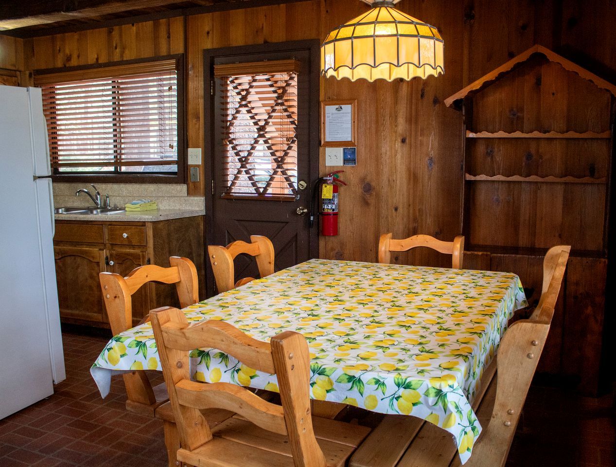 A wooden cabin interior with a dining table set for four, a kitchen area, and a window with blinds.