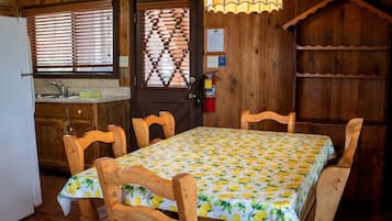 A wooden cabin interior with a dining table set for four, a kitchen area, and a window with blinds.
