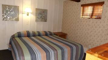 Une chambre à coucher avec une couvre-lit à rayures, deux lampes fixées au mur, une fenêtre avec des stores et une table de chevet en bois.