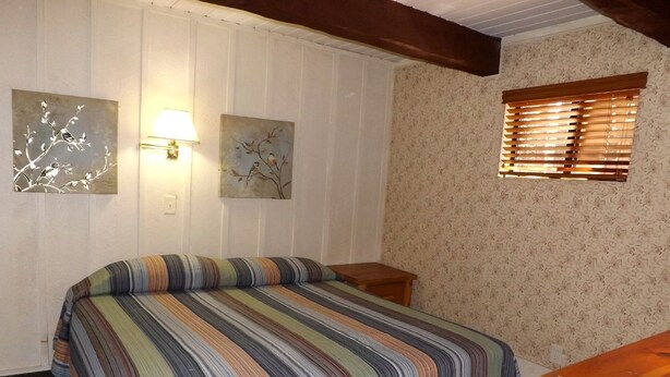 A bedroom with a striped bedspread, two wall-mounted lamps, a window with blinds, and a wooden nightstand. - Spacious Cabin with Large Porch Ideal for Groups in Arizona (Flagstaff)