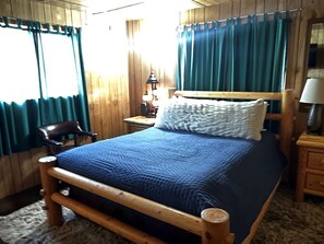 A wooden-framed bed with a blue quilt, a chair, a nightstand with a lamp, and a window with curtains. - Relaxing Log Cabin Nestled in the Trees near Yellowstone, Idaho (Island Park)
