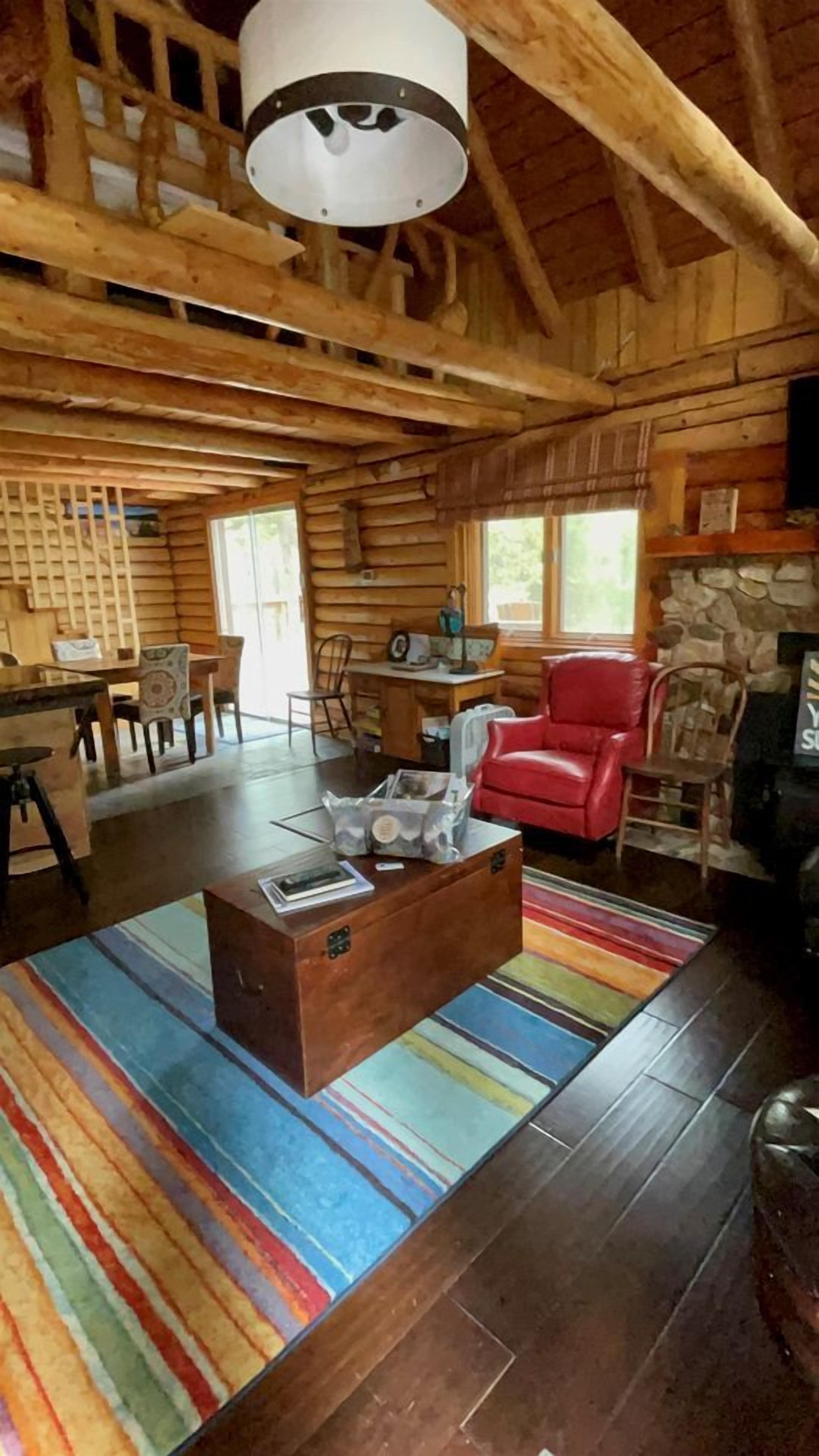 A log cabin interior with a wooden ceiling, a red leather sofa, a stone fireplace, and a colorful striped rug.