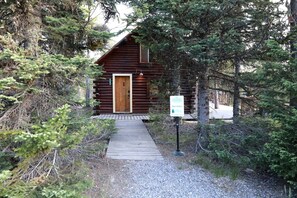 A log cabin with a wooden door, a gravel path leading to it, and a signboard in the foreground. - Relaxing Log Cabin Nestled in the Trees near Yellowstone, Idaho (Island Park)