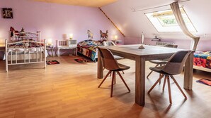 A room with a wooden floor, a bed with a colorful quilt, a dining table with chairs, a window with curtains, and a small table with a lamp. - Farmhouse Vacation Rental for 15 People Near the Somme Bay (Friville-Escarbotin)