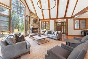 A spacious living room with large windows, a stone fireplace, and a cozy seating area. - Monthly Lease - Signature Lodge at Martis Camp by Tahoe Getaways - 4BR Estate! (Truckee)