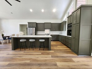 A modern kitchen with a central island, bar stools, and dark cabinetry. - Brand New Family Lake Retreat with Two Living Areas (Greers Ferry)