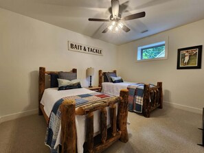 A bedroom with two wooden beds, a ceiling fan, a window with a view, and a wall sign reading 'BAIT & TACKLE'. - Brand New Family Lake Retreat with Two Living Areas (Greers Ferry)