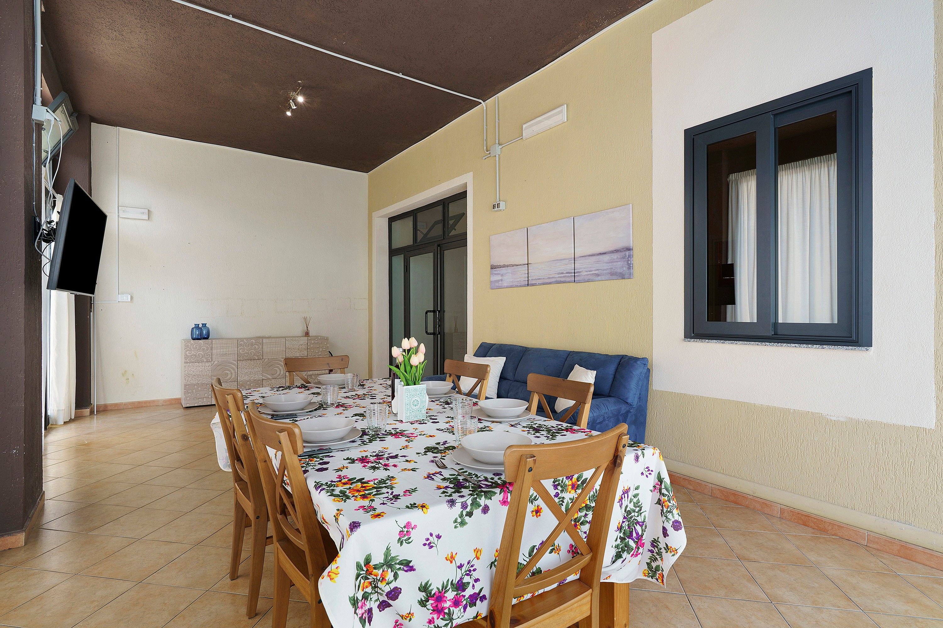 A dining area with a table set for four, surrounded by wooden chairs, a blue sofa, and a window with curtains.