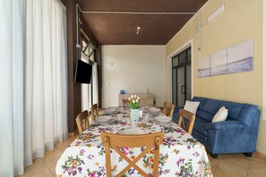 Zona de dinar amb taula per a quatre, cadires, un sofà blau i un televisor penjat a la paret.