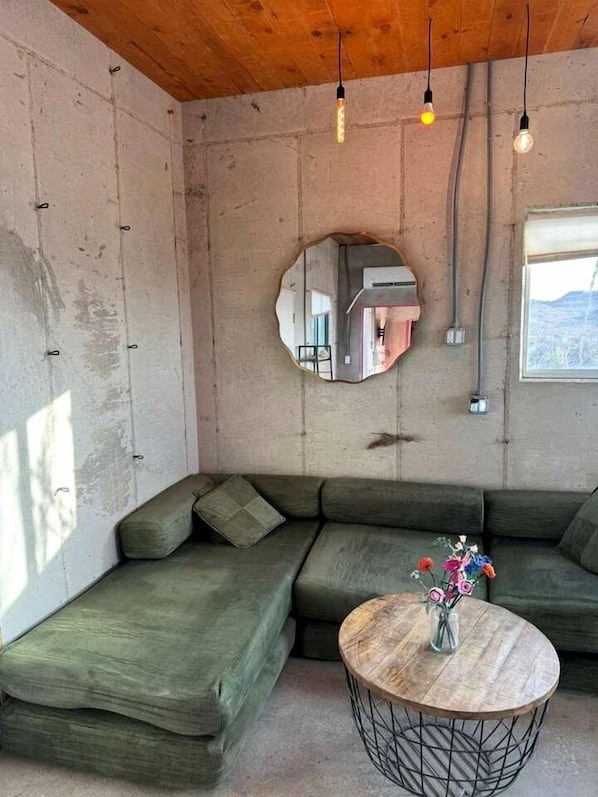Una habitación con un sofá seccional verde, una mesa de centro redonda de madera, un espejo circular e iluminación colgante.