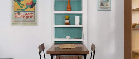 A dining area with a wooden table and chairs, a teal shelf unit with decorative items, and two framed posters on the wall.
