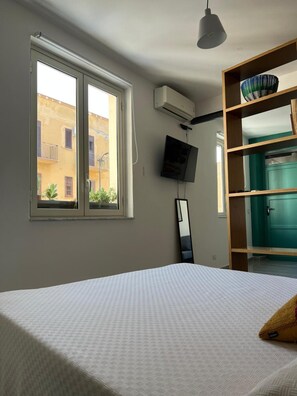 Ein Schlafzimmer mit einem Bett, einem an der Wand befestigten Fernseher und einem Fenster mit Blick auf Gebäude.
