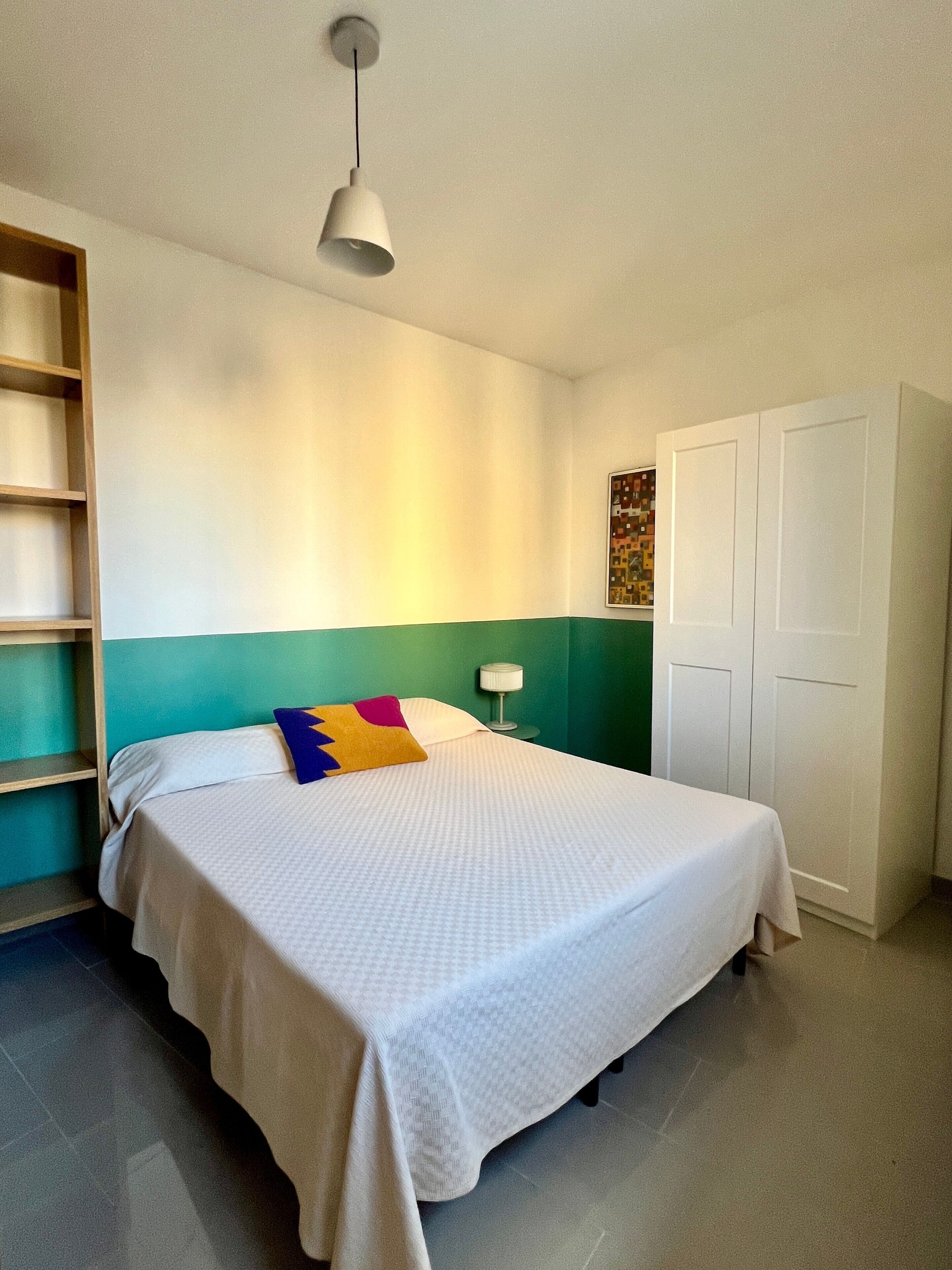 A single bed with a white bedspread, a colorful pillow, a bedside lamp, and a wardrobe in a room with teal and white walls.