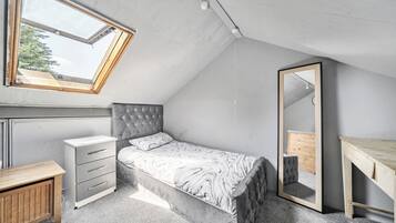 Un dormitorio con techo inclinado, una cama con cabecera capitonada, una mesita de noche, un aparador, un espejo y un tragaluz.